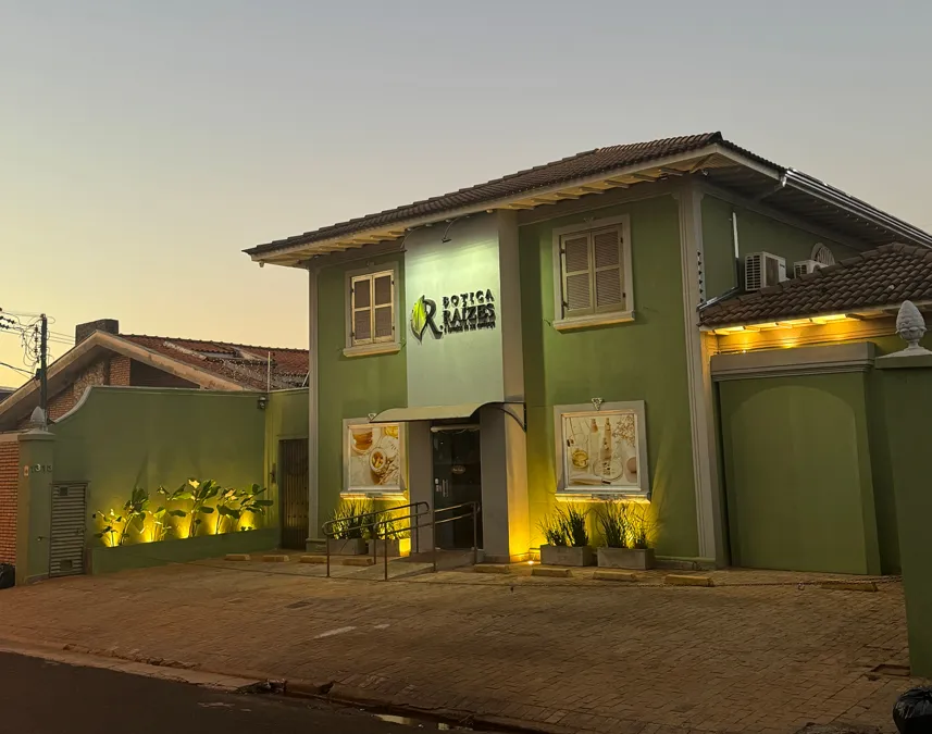 Fachada da Botica Raízes, farmácia de manipulação com identidade visual verde, iluminação externa e jardim decorativo.